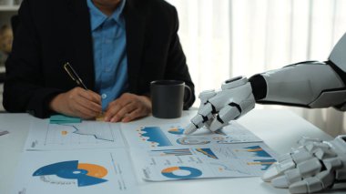 Bir insan ve robotik kol bir iş toplantısında işbirliği yapar, finansal raporları analiz eder ve modern ofis ortamında veri içgörülerini tartışır. Noogenesis