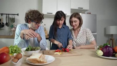 Çekici beyaz anne ve baba Asyalı kızıyla taze yemek pişirmek için birlikte vakit geçiriyorlar. Enerjik anne ve baba mutfakta salata yapmak için taze sebze hazırlıyor. Sağlıklı yiyecekler. Pedagoji.