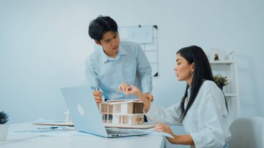 Profesyonel Asyalı erkek mimar ev modelinin uzunluğunu ölçmek için cetvel kullanırken, beyaz tenli genç meslektaşı ev modeliyle toplantı masasındaki verileri analiz etmek için dizüstü bilgisayar kullanıyor. Tertemiz..
