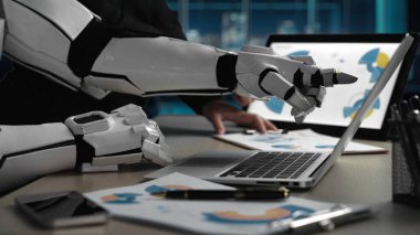 Gelecekçi bir robot el, modern ofis ortamında dizüstü bilgisayarla etkileşime girer, veri ve grafikleri analiz eder, yapay zeka teknolojisinin iş dünyasıyla birleşimini gösterir. Noogenesis
