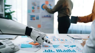 Veri sunumları ve grafikleri analiz eden, yenilikçi bir çalışma alanında teknoloji ve profesyoneller arasındaki işbirliğini vurgulayan robotik bir kolu gösteren gelecekçi bir ofis sahnesi. Noogenesis