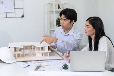 Profesyonel Asyalı erkek mimar ev modelinin uzunluğunu ölçmek için cetvel kullanırken, beyaz tenli genç meslektaşı ev modeliyle toplantı masasındaki verileri analiz etmek için dizüstü bilgisayar kullanıyor. Tertemiz..