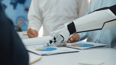 Gelecekçi bir robot kolu, takım üyeleri modern bir ofis ortamında işbirliği yaparken, çizelge ve grafikleri analiz eden bir iş toplantısındadır.