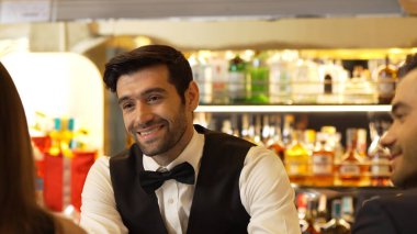 Güzel görünümlü barmen, içki tezgahındaki çiftle samimi bir gülümseme sohbeti yaparken aynı zamanda hoş geldin içkisi ve özel menü listesi öneren çekici profesyonellik. Vinosity.