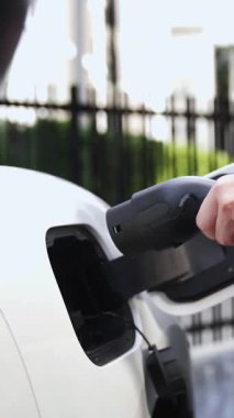İş kadını akıllı EV şarj aleti takıyor ve şehir otoparkındaki EV şarj istasyonundan elektrikli araba şarj ediyor. Geleceksel sürdürülebilir güç ve EV araba kullanımı. İnceleyin
