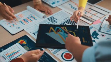 Stratejik bir toplantıya katılan bir grup profesyonel, tabloya yayılmış tablolar ve grafikler üzerindeki veri analizlerini tartışıyor, işbirliği ve verimliliği gösteriyor. SACTR