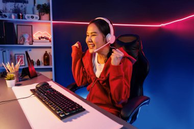 Yüzünde kulaklık renginde kulaklık takan, neon odada online oyun oynayan, Esport meziyetli takım oyuncularına yumruk kaldırarak oynayan güzel Asyalı kız flamasının sunucu kanalı. Taktik.