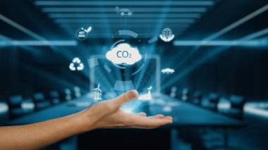 Karbon ayak izi azalması, rüzgar enerjisi ve geri dönüşüm de dahil olmak üzere sürdürülebilirlik kavramlarını temsil eden dijital simgelerin yer aldığı modern bir ofise uzanan bir el. Auvana