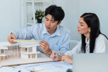 Profesyonel Asyalı erkek mimar ev modelinin uzunluğunu ölçmek için cetvel kullanırken, beyaz tenli genç meslektaşı ev modeliyle toplantı masasındaki verileri analiz etmek için dizüstü bilgisayar kullanıyor. Tertemiz..