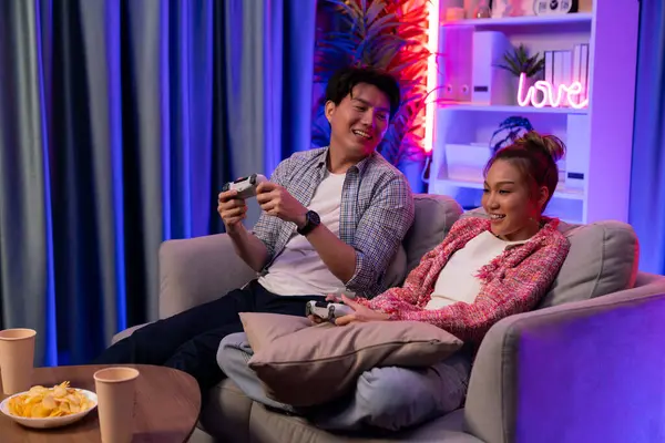 Mutlu çiftin oturduğu kanepe joysticklerle video oyunu oynuyor, meydan okuyan gülümseyen adam, evdeki modern neon ışıklı oturma odasında kazanmak için yumruğunu kaldırıyor. İnfobahn.