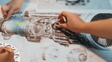 Endüstriyel bir motoru gösteren detaylı mekanik planların yakın görüntüsü. Eller, bir tasarım atölyesinde çalışabilecek bileşenleri dikkatlice inceleyerek hassasiyeti vurguluyor. SACTR