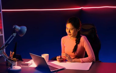Geceleri sosyal medyada canlı yayında pozitif podcastla konuşan gülümseyen genç, güzel Asyalı etkileyici pembe neon ışıklı modern stüdyoda sunum için dizüstü bilgisayar kullanıyor. Taktik.
