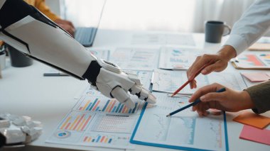 Bir robot kol ve insan eli modern bir ofiste iş verilerini ve grafiklerini analiz ederek teknoloji ve insan uzmanlığının kaynaşmasını gösteriyor. Noogenesis