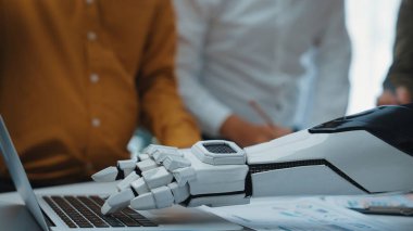 Gelecekçi bir robotik el, modern ofis ortamında dizüstü bilgisayarla etkileşime girer ve iş yerindeki teknoloji ile insan işbirliğinin karışımını vurgular. Noogenesis