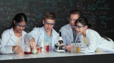 Çok kültürlü bir öğrenci mikroskop altında incelerken güzel bir kız da numuneyi inceliyor. BTMM sınıfında karatahta bilim teorisi deneyi yapan liseli çocuklara bakan öğretmen. Öğretim