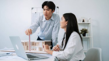 Profesyonel Asyalı erkek mimar ev modelinin uzunluğunu ölçmek için cetvel kullanırken, beyaz tenli genç meslektaşı ev modeliyle toplantı masasındaki verileri analiz etmek için dizüstü bilgisayar kullanıyor. Tertemiz..