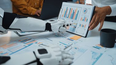 Bir insan iş dünyasındaki yenilikleri ve takım çalışmalarını ön plana çıkarırken, modern ofis sahnesinde robot kolu veri tablolarıyla etkileşime giriyor. Noogenesis