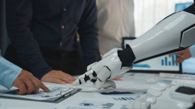 Robot bir elin insan elini sıktığını gösteren, takım çalışması ve teknoloji entegrasyonunu gösteren modern bir iş toplantısı. Belgeler ve çizelgeler mevcut, veri analizleri vurgulanıyor. Noogenesis