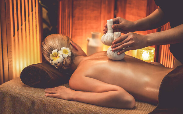 Hot herbal ball spa massage body treatment, masseur gently compresses herb bag on woman body. Tranquil and serenity of aromatherapy recreation in warm lighting of candles at spa salon. Quiescent