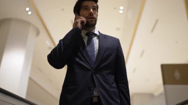 Profesyonel bir iş adamı yürüyen merdiven kullanırken telefonla konuşuyor. Beyaz tenli yönetici, pazarlama fikri ya da planı rapor etmek ve tartışmak için iş ekibiyle konuşuyor. Ön manzara. Sevinçli..