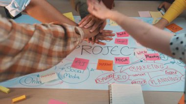 Çeşitli eller renkli bir proje planı üzerinde bir araya gelerek, pazarlama ve içerik stratejisindeki işbirliğini vurgulayarak, etkili takım çalışması ve planlama araçlarını sergileyerek. SACTR