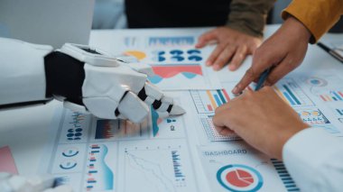 Bir robot el, iş haritaları ve grafiklerle dolu bir masada insan eliyle etkileşime girer. Ofis ortamında teknoloji ve takım çalışması harmanlanır. Noogenesis