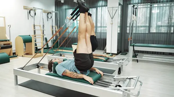 Müşteri, spor salonundaki yenilikçi bir makinede kayışla ters bacak kaldırma hareketi yapıyor. Çekirdek bağlantısını, dengesini ve esnekliğini geliştirmek için ters takla atan iki mutlu antrenör. Alışkanlık.