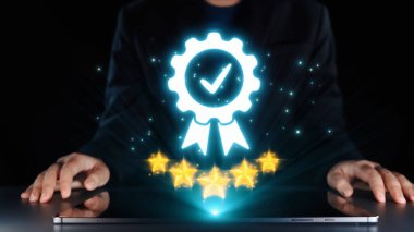 Koyu renk takım elbiseli bir kişi, dijital rozeti parlayan bir dizüstü bilgisayarın önünde duruyor ve beş parlayan yıldız, karanlık bir arkaplanda kalite ve başarıyı simgeliyor. Dörtgen