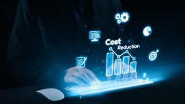 Klavyede oturan profesyonel bir birey, modern bir çalışma ortamında maliyet azaltma ve finansal analiz temalarını gösteren grafikler ve simgelerin holografik görüntüleriyle etkileşim halindedir. Dörtgen