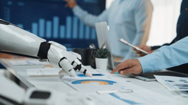 Modern bir ofis sahnesinde robot bir el ile veri analizi yapan ve profesyonel bir çalışma alanında teknolojiyi sergileyen insan meslektaşları arasındaki işbirliği vurgulanır.