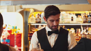 Güzel görünümlü barmen, içki tezgahındaki çiftle samimi bir gülümseme sohbeti yaparken aynı zamanda hoş geldin içkisi ve özel menü listesi öneren çekici profesyonellik. Vinosity.