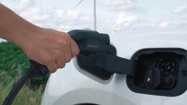 Elektrikli arabasıyla ilerici bir adam, EV arabası gelecekteki sürdürülebilir enerji kavramı olarak rüzgar türbiniyle yeşil alandaki enerji şarj etme istasyonundan enerji yüklüyor. Enerji üreteci olan elektrikli bir araç..