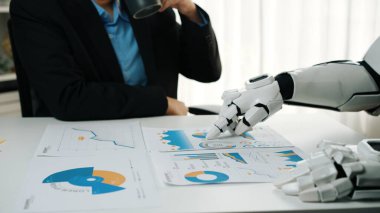 Bir insan ve bir robot modern bir çalışma alanında işbirliği yapıyor. Robot veri tablolarına işaret ederken, insan kahveden zevk alıyor. Analizde gelecekteki işbirliğini simgeliyor. Noogenesis