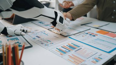 Robot bir kol, modern bir ofiste işbirliğini sembolize eden bir insanla tokalaşır. Takım çalışmaları chartlar tarafından temsil edilen veri odaklı analizler üzerine yoğunlaşır. Noogenesis