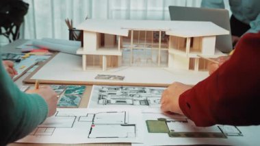 İşbirlikçi mimari tasarım oturumunda el çizimleri ve bir model üzerinde çalışırken görülüyor. Parlak çalışma alanı planlar ve malzemelerle dolu. SACTR