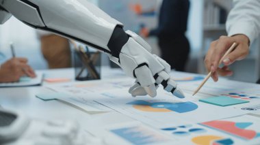 Robotik bir el modern ofis ortamında iş grafiklerine uzanıyor, çeşitli profesyoneller arasında bilgilendirilmiş kararlar için verileri analiz eden bir işbirliği sergiliyor.