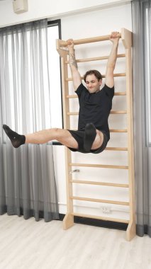 Asılı bacak egzersizi yapan mutlu bir adam vücut kaslarını güçlendirmek ve beden eğitimini iyileştirmek için duvar parmaklıkları üzerinde egzersiz yapıyor. Alt karın bölgesini iyileştirmek için erkek antrenör bacak yükseltme ameliyatı. Alışkanlık.