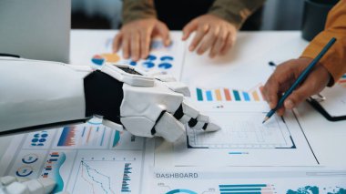 Gelecekçi bir robot kolu modern çalışma masasında çeşitli grafik ve grafiklerle veri analizinde profesyonellere yardımcı olur, teknoloji ve işbirliğinde yenilik sergiler.