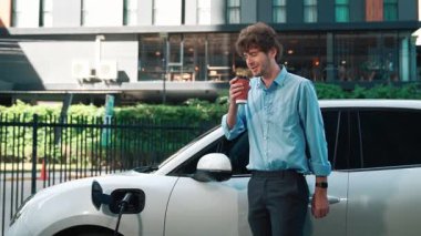 Takım elbiseli, hırslı bir işadamı elektrik kablosu ve yenilenebilir enerji enerjili bir elektrikli araçla şarj istasyonunda dururken elektrikli aracına yaslanıyor..