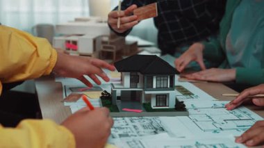 Bir grup profesyonel, takım çalışması ve yaratıcılığı vurgulayarak, modern bir çalışma alanında minyatür modeller ve ayrıntılı kat planları kullanarak mimari planlamada işbirliği yapmaktadır. SACTR