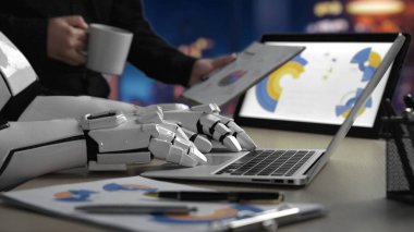 Robot bir el, gece şık bir ofiste dizüstü bilgisayarla ve belgelerle etkileşime girer. Teknoloji odaklı bir iş ortamında çalışmanın geleceğini ön plana çıkarır. Noogenesis