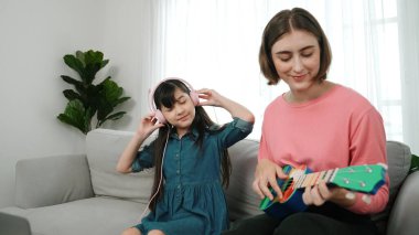 Asyalı tatlı kız kulaklıkla müzik dinlerken güzel anne de akustik müzik öğretirken ukulele çalıyor. Sevimli çocuk enstrüman öğreniyor. Pedagoji.