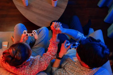 Çarpışan joystickleri olan, el ele tutuşarak kazanan, modern evdeki neon ışıklı oturma odasının arka tarafında video oyunu oynayan genç çift oyuncusunun en iyi görünümü. İnfobahn.