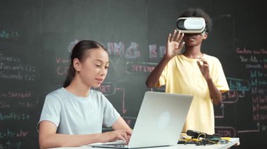 Genç ve güzel akademik öğrenci yazma, kodlama ve programlama sistemini bilgisayarı ya da aygıtı olan masaya yerleştirirken Afrikalı çocuk metaevrene girmek için kulaklık takıyor. yenilikçi eğitim. Öğretim