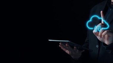 Bir kişi, bulut bilgisayar simgeleri ve dijital sembollerle çevriliyken, veri transferi, güvenlik ve modern teknoloji kavramlarını karanlık bir ortamda gösteren bir tablet tutuyor. Dörtgen