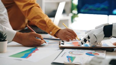 İnsan eli, modern ofis ortamında çizelge ve verileri analiz etmek için robot kolla işbirliği yapar. Teknoloji ve takım çalışmasının iş etkinliklerindeki karışımını gösterir. Noogenesis