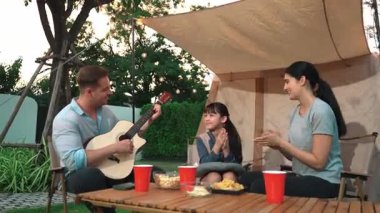 Baba gitar çalarken anne ve kız birlikte kampta oturuyorlar. Ebeveynler, genç nesil çapraz kuşak farklılıklarıyla zaman geçirmek için açık hava kamp aktivitelerini kullanıyorlar. Uyumsuzluk.