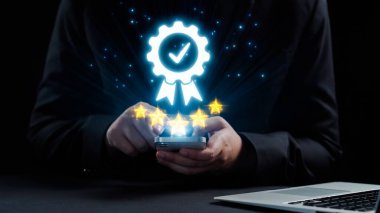 Bir kişi, yenilikle dolu profesyonel bir atmosferde başarı ve tanınmayı temsil eden parıldayan bir ödül sembolü ve yıldızlarla süslenmiş bir akıllı telefon tutar. Dörtgen