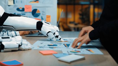 Modern bir ofis sahnesinde, bir robot el veri analizinde insan eliyle işbirliği yapıyor. İş haritaları ve teknoloji araçları yenilik ve takım çalışmasını teşvik ediyor. Yaratıcılık