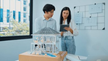Güzel, genç, beyaz bir proje yöneticisi Asyalı mühendise, mühendislik masasında ev modelini ölçerken tablet kullanarak bina detaylarını anlattı. Tertemiz.